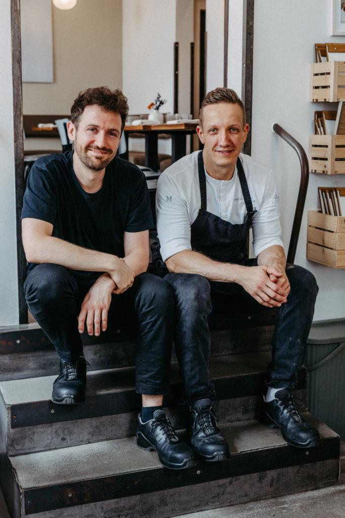 Zwei Mitarbeiter des Willichs Restaurants sitzen auf einer Treppe und lächeln in die Kamera – authentisches Teamfoto mit Wohlfühlatmosphäre